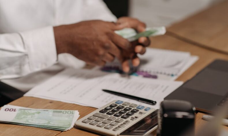 Hands counting euro bills on a wooden desk with calculator, financial documents, and laptop nearby. - Photo by Pavel Danilyuk on Pexels
