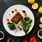 Top view of delicious fried fish served in white plate with fresh salad and spinach and placed on black table table with lemons tomatoes and sauces - Photo by Ray Piedra on Pexels