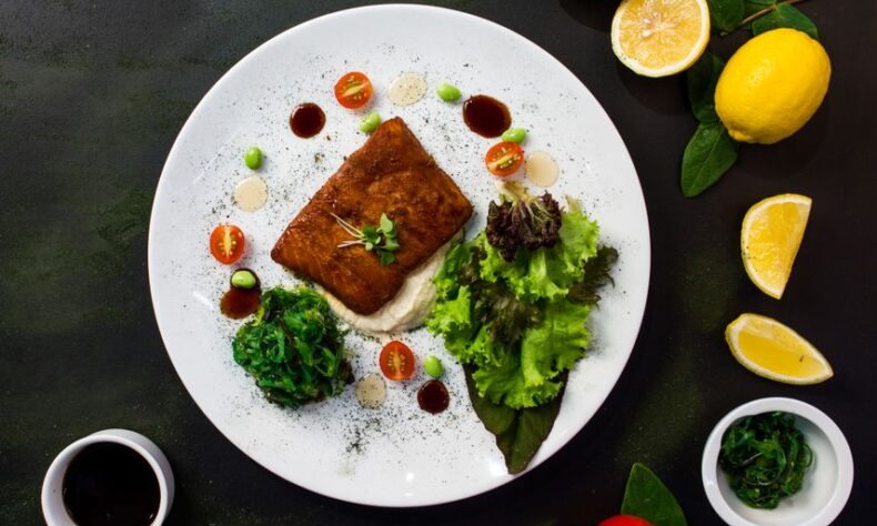 Top view of delicious fried fish served in white plate with fresh salad and spinach and placed on black table table with lemons tomatoes and sauces - Photo by Ray Piedra on Pexels